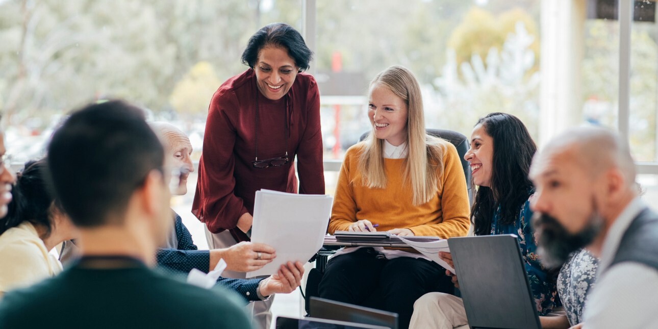 Eight smiling colleagues of different ages, genders and ethnicities gathered around a coffee table in discussion and looking at paperwork.