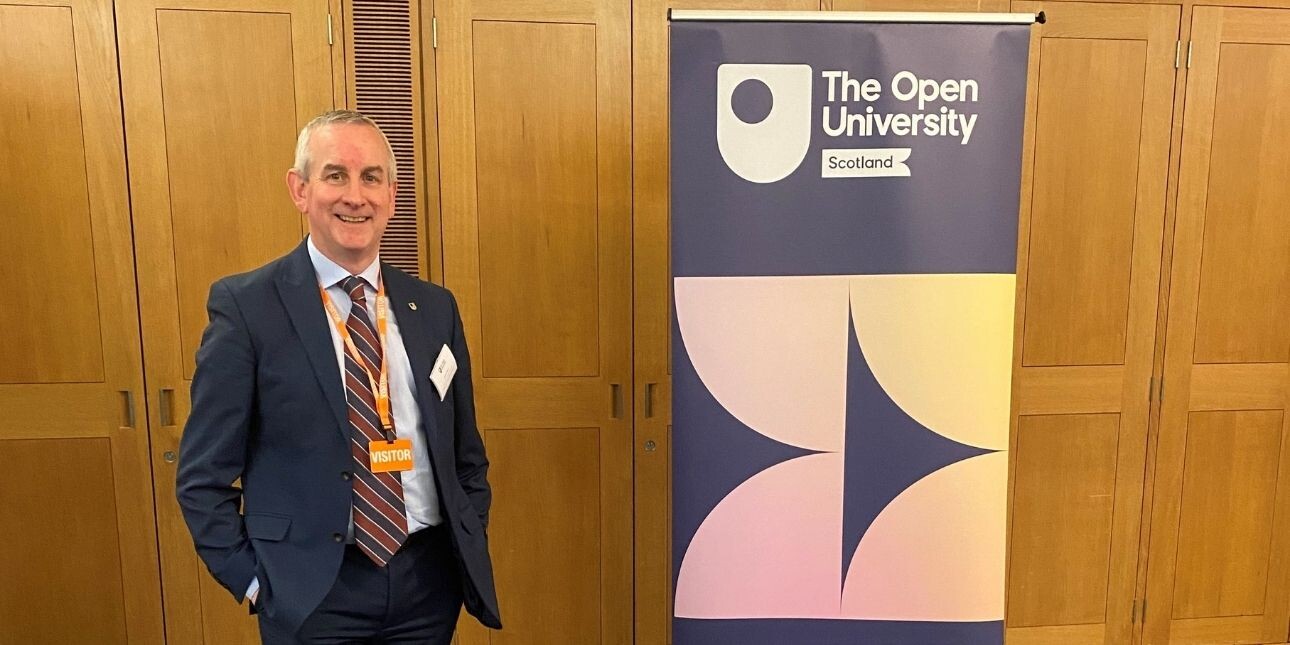 A smiling Keith Robson stood in front of a wood panelled wall. Keith is a white man with grey hair, who wears a navy suit. To his is left is a banner featuring the words Open University Scotland.