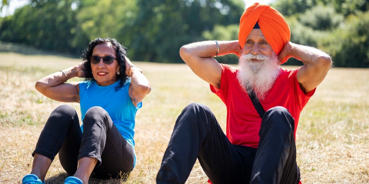 An elderly Asian couple perform sit ups on the grass. On the left is a woman wearing a blue vest and dark leggings. Next to her is a man wearing a red t-shirt, orange vest and blue tracksuit trousers.