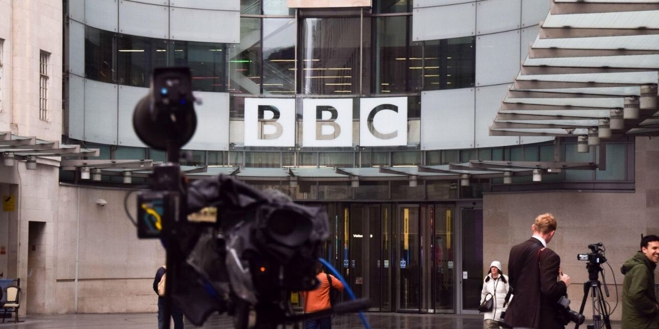 Broadcasting equipment and reporters outside the glass fronted Broadcasting House. The BBC logo is positioned in the background.