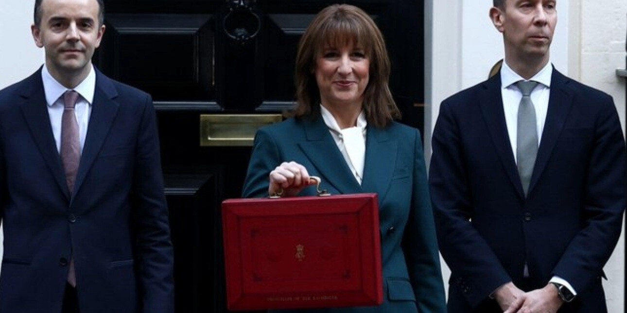 Chancellor Rachel Reeves flanked by two colleagues hold up a red briefcase outside the black door of 11 Downing Street. Reeves is a white woman with bobbed brown hair, who wears a navy suit.