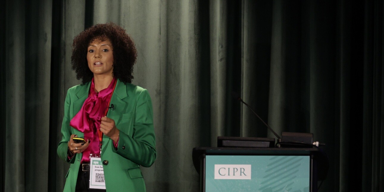 Naomi Goodman on the left of the stage. She is wearing a green jacket over a bright pink blouse.