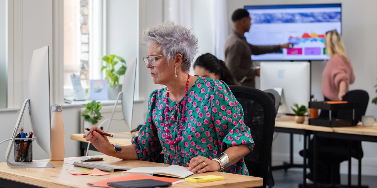 A colour photo of a middle-aged white woman with grey hair, glasses and a turquoise and pink dress sat at her desk in an office with paperwork and a monitor in front of her.