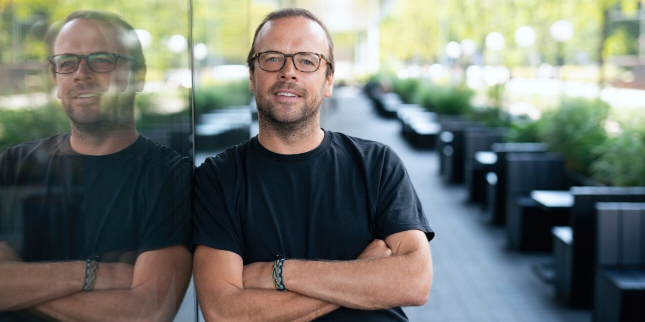 A colour portrait of James Peach leaning against a large glass window which shows his reflection. James is a white man with short dark hair and beard, wearing glasses and a black top.