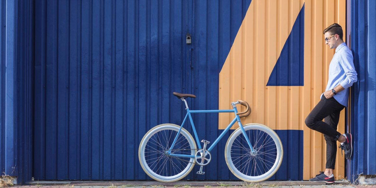 A turquoise bike with white wheel trims stood against blue corrugated doors which feature a yellow painted four numeral. To the right, a white man wearing denim stands side on looking at the bike.