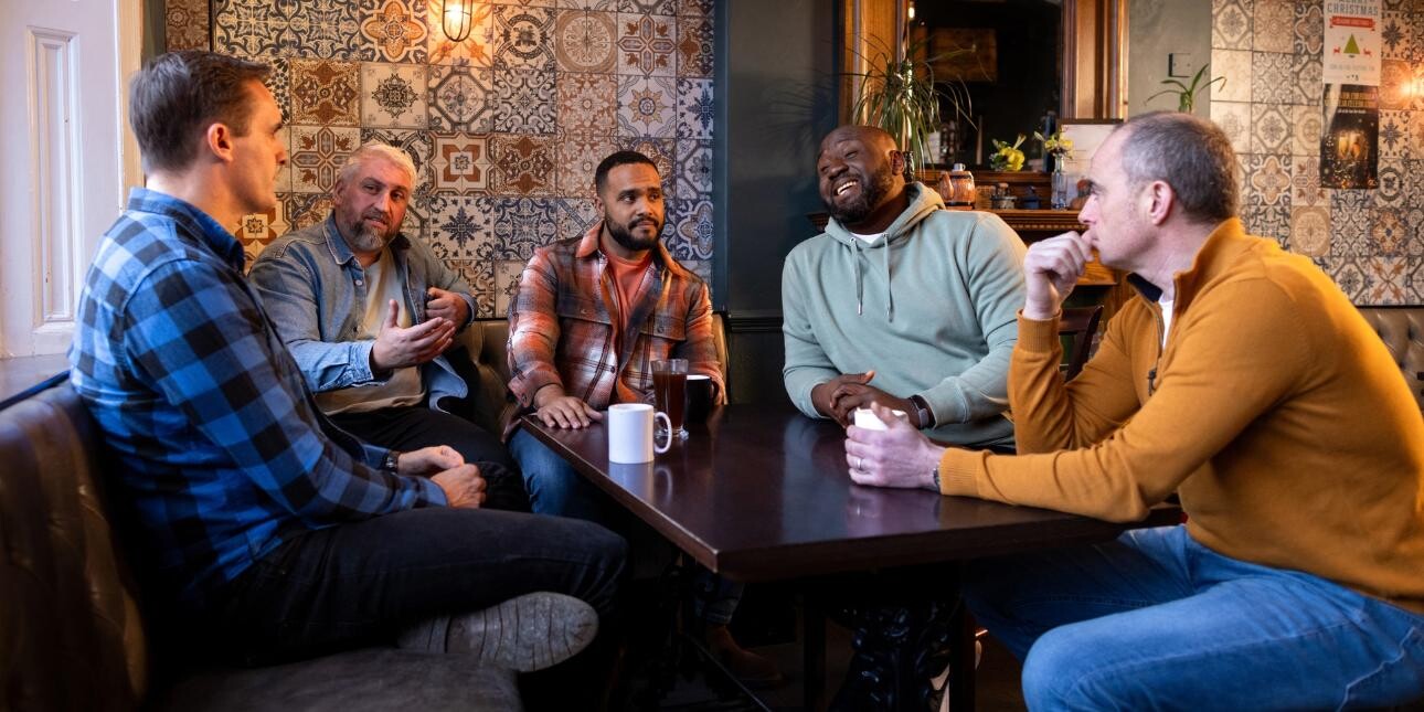 Five middle aged men sat chatting around a dark wooden table in a pub or cafe. The wall behind them features colourful, decorative tiles and a large mirror.