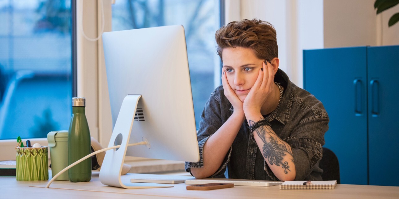 A white woman with short brown hair and wearing denim shirt rests her elbows on the desk and her head in her hands as she looks at her computer screen. She is in office with a window and assorted belongings on desk.