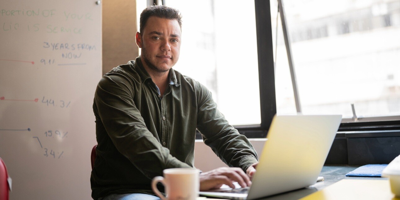 A transgender mature man with dark hair, who wears a dark green shirt, sat at a desk using a laptop in an office next to a window.