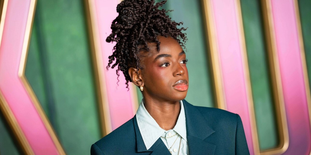A close up portrait of Cat Burns wearing a dark suit in front of a pink and green hoarding.
