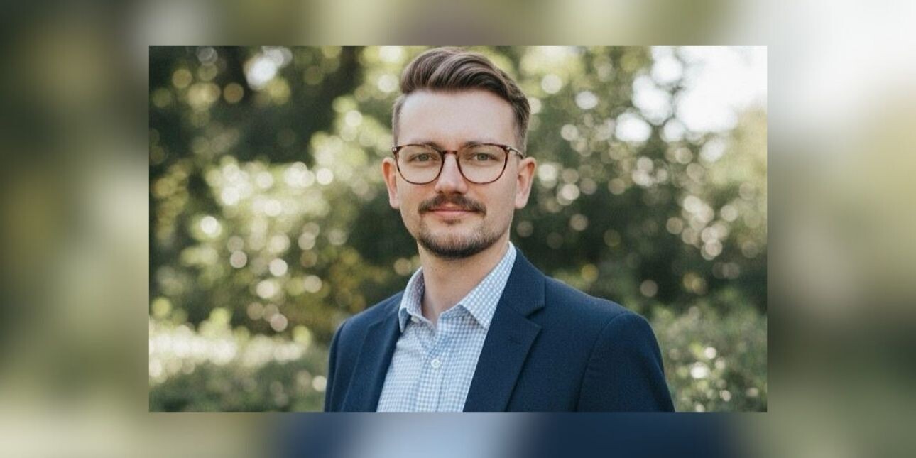 A colour portrait of James Sharp in front of trees.Sharp is a white man with dark hair who wears glasses and a grey suit jacket.