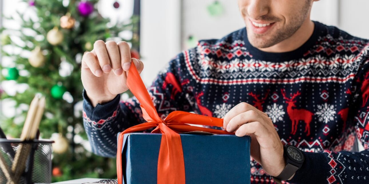 A close up of a white man wearing a Christmas jumper, sat at an office desk untying an orange ribbon wrapped around a gift wrapped in navy paper.