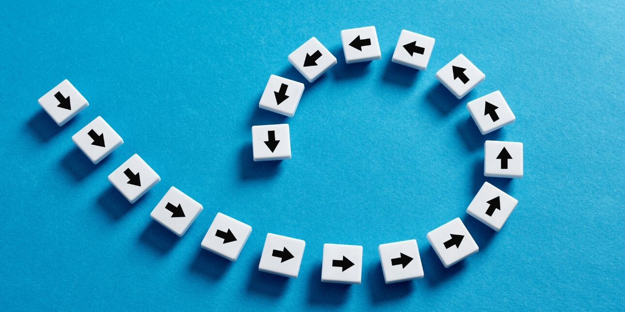 Concept of being stuck in a circle. A line of white tiles with black arrows are placed on a blue surface. The line loops round on itself.
