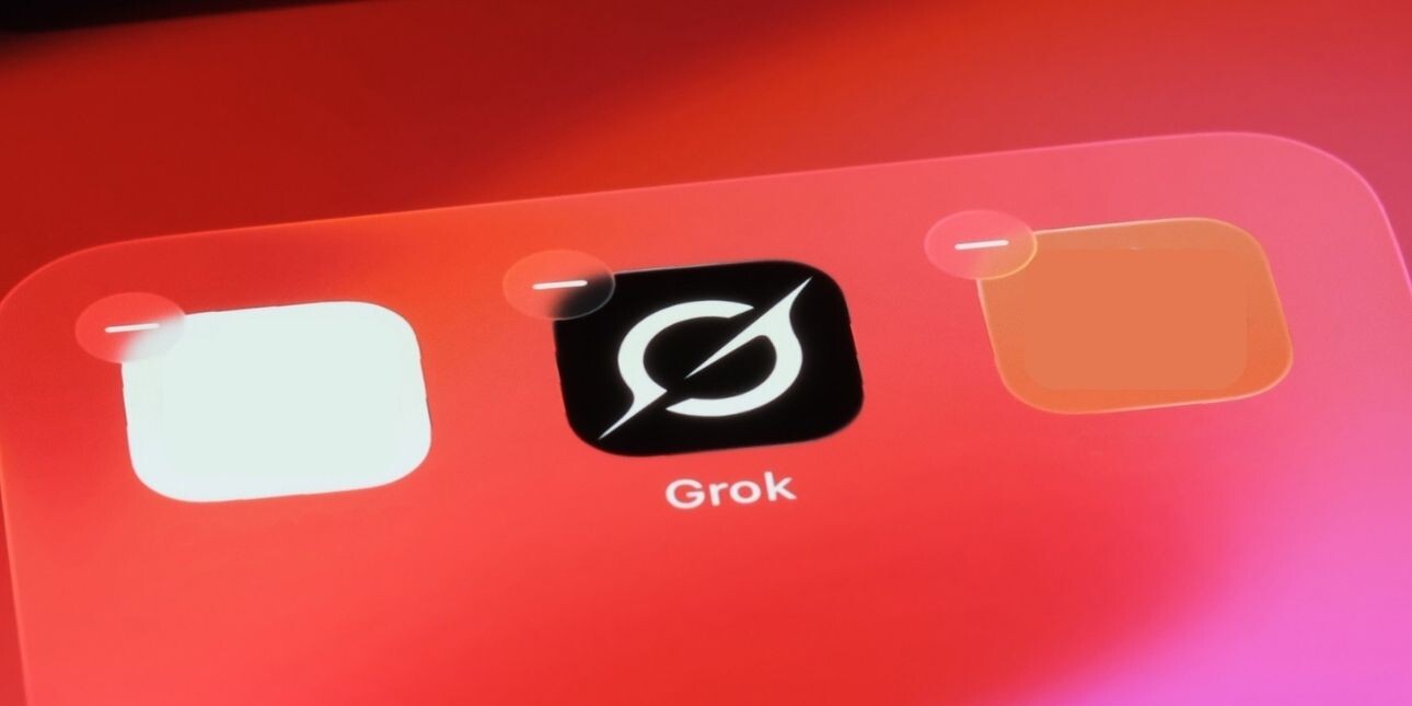 A photo of the Grok app on a pink-orange mobile phone screen. The Grok logo is a white stylised uppercase G on a black background.