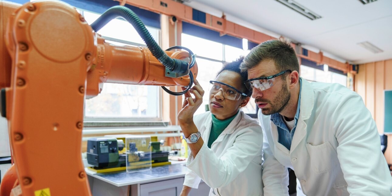 Two colleagues in white lab coats and protective glasses - a Black woman on the left and a white man on the right - inspect an orange robotic arm in a laboratory.