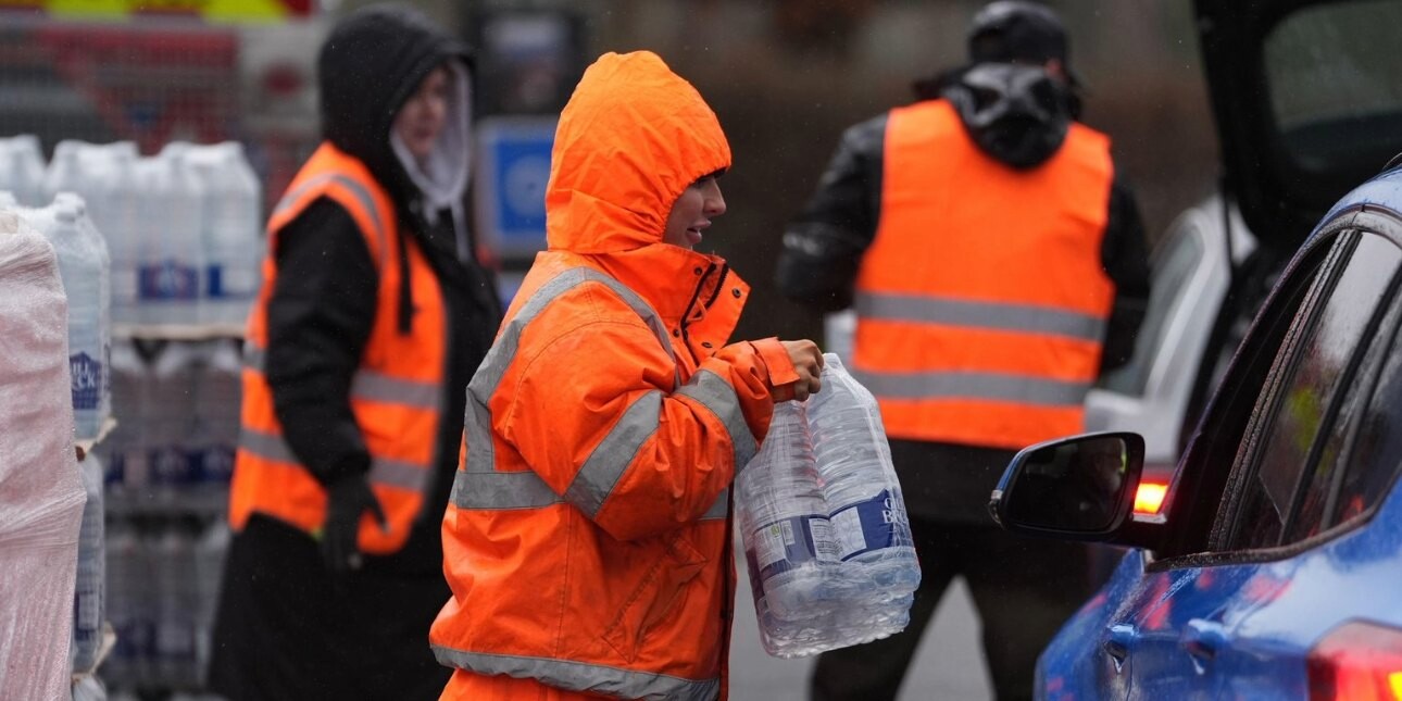 A worker in orange hi-vis clothing hands over bottled water to unseen recipients of a blue car.