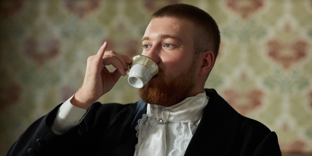 A white man with cropped brown hair and wearing a formal dark jacket and white ruffled shirt holds a porcelain tea cup with his pinkie finger extended.