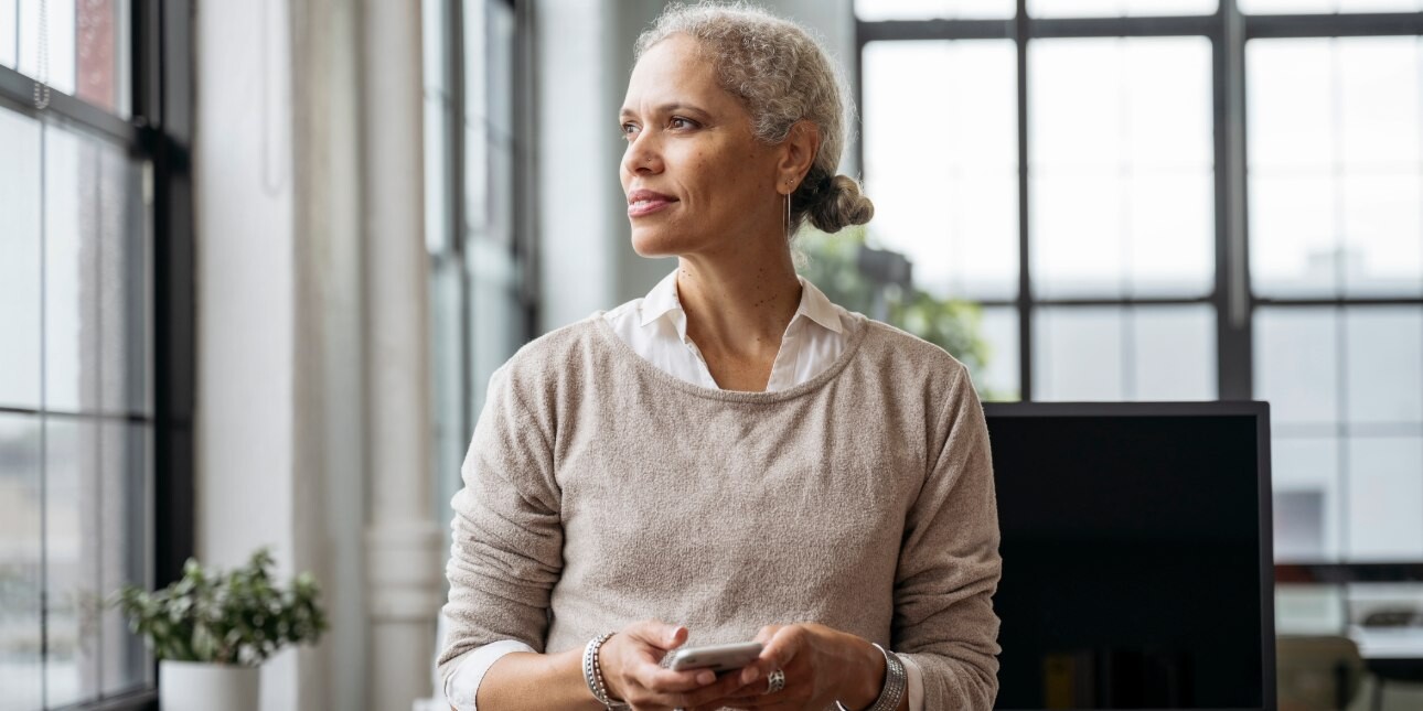 In a light-filled office, a contemplative businesswoman stands with her head faced towards the window while holding a phone in her hand.