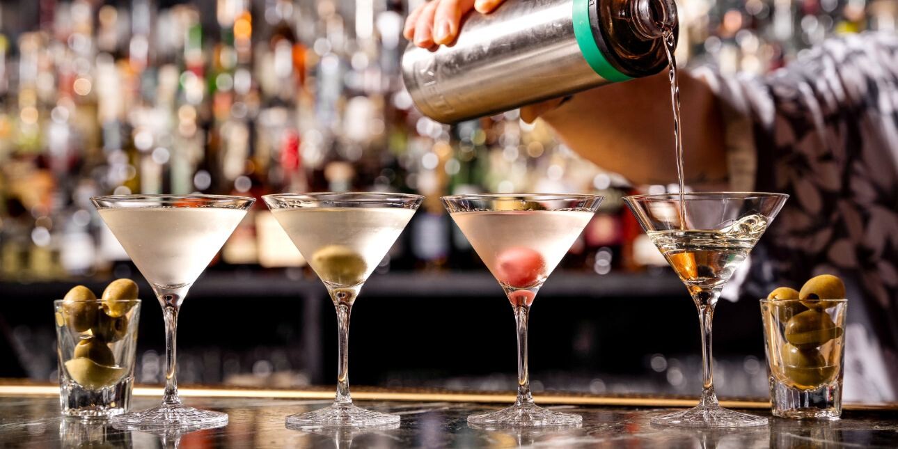 Four glasses of martini on a bar flanked by two small glasses of olives. An unidentified mixologist pour the drink from a silver shaker into the fourth martini glass. The background is out of focus.