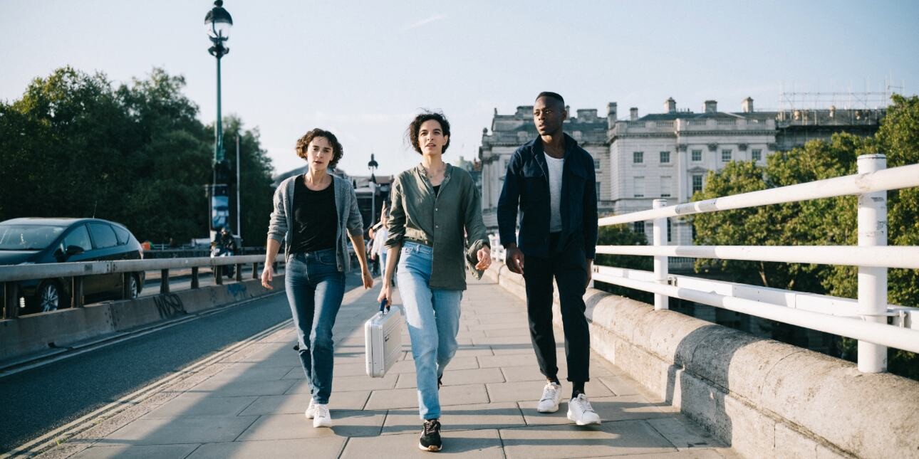 Three people - two women and one man - striding across a bridge in London. The woman in the middle is holding a metallic briefcase.
