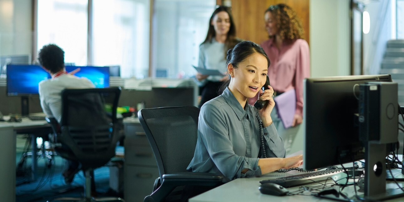 A woman sat behind her desk speaks on the phone in an office. Two other woman are stood talking behind her, while the back of a man also sat at desk can be seen to the left.