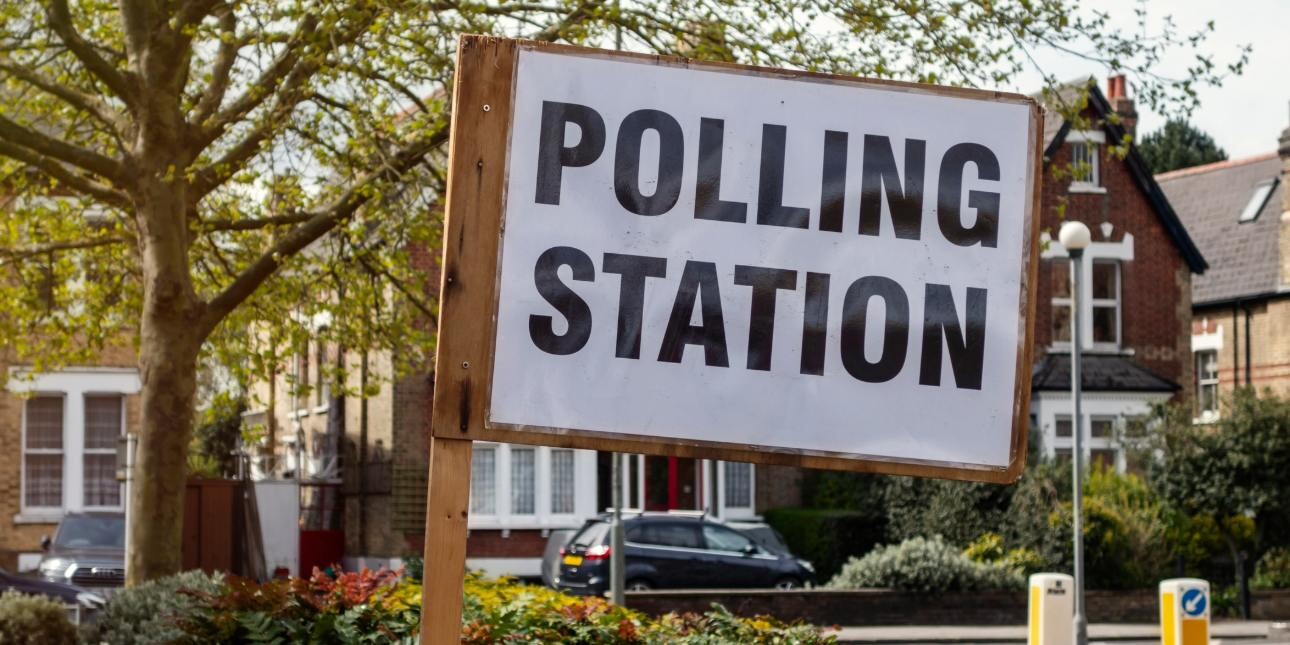 A polling station flag consisting of black text on a white background upon a wooden post. In the background are three-storey British red-brick houses and trees.