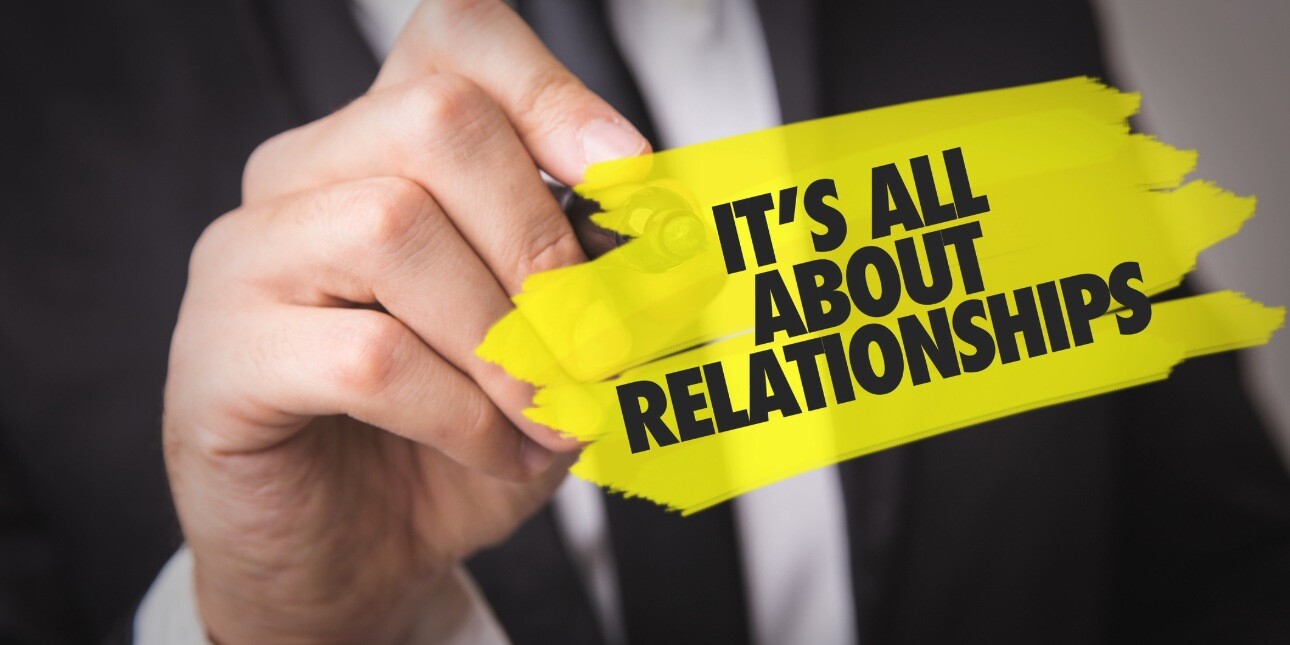 A close up of the hand of a white male wearing a black suit. He is holding a pen to a transparent board. Superimposed in black writing on a yellow background are the words: it's all about relationships.