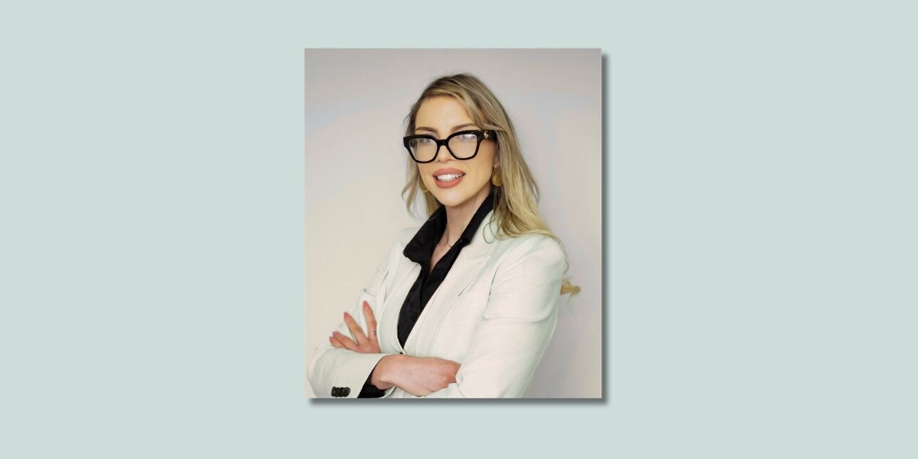 On a grey background, a colour portrait of Amy Simpson. Amy is a white woman with long blond hair who is wearing a light coloured jacket.