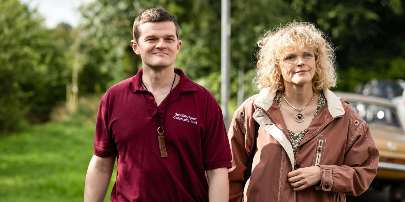 A still from the film I Swear, with actors Robert Aramayo and Maxine Peak walking along a suburban street with trees in the background.