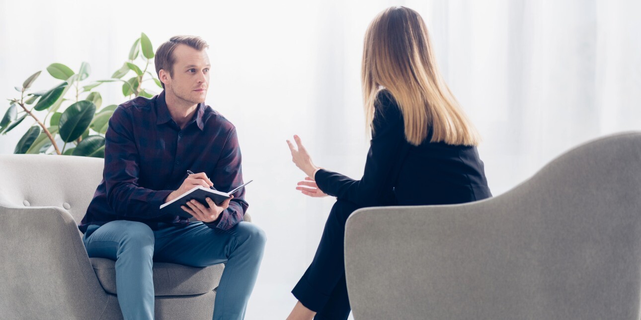 In a light filled room, a causally dressed white male journalist holds a notebook while interviewing smartly dressed white business woman with blond hair. The photo is taken from over her shoulder.