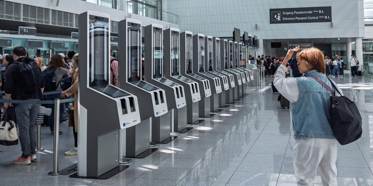 The back of a person wearing a blue top and white trousers looking at a row of tall, grey coloured computer scanners in a spacious airport.