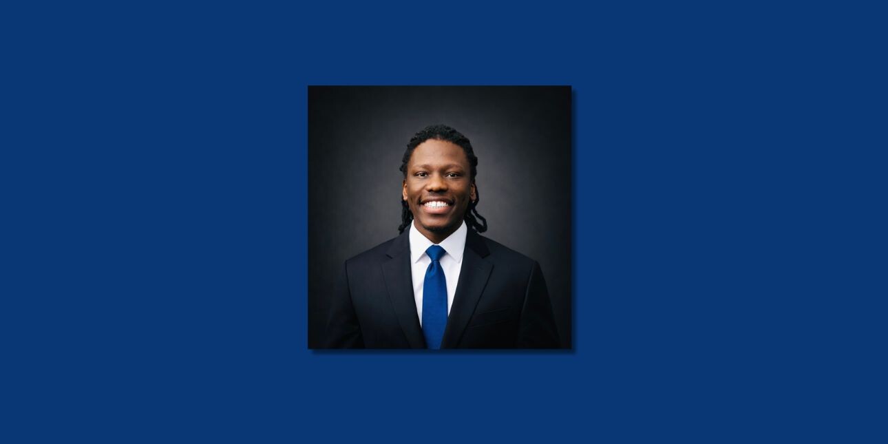 A colour portrait on a blue background of Emmanuel Kisolom smiling. Emmanuel is a Black man who is wearing a dark suit and a navy tie.