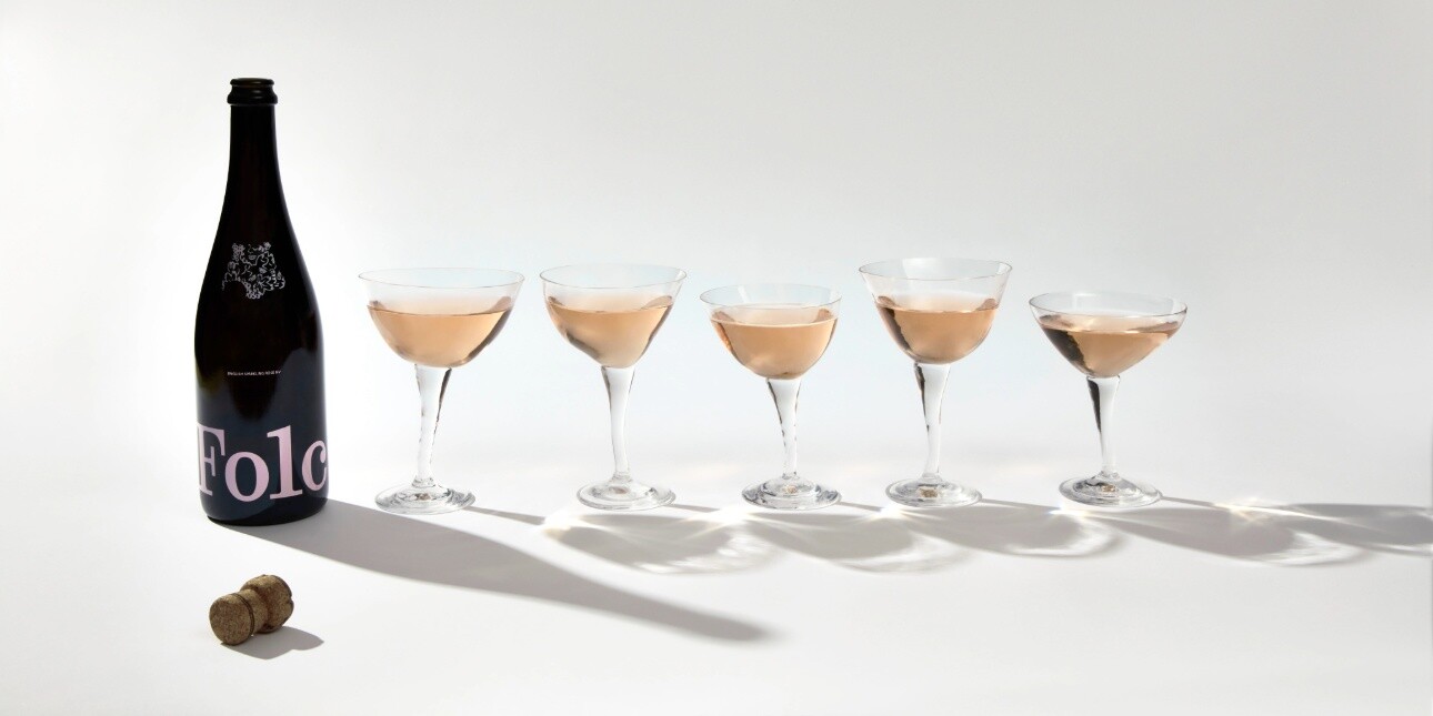 A group shot on a white background of a dark uncorked Folc wine bottle alongside five different-shaped stem glasses of sparkling rosé wine
