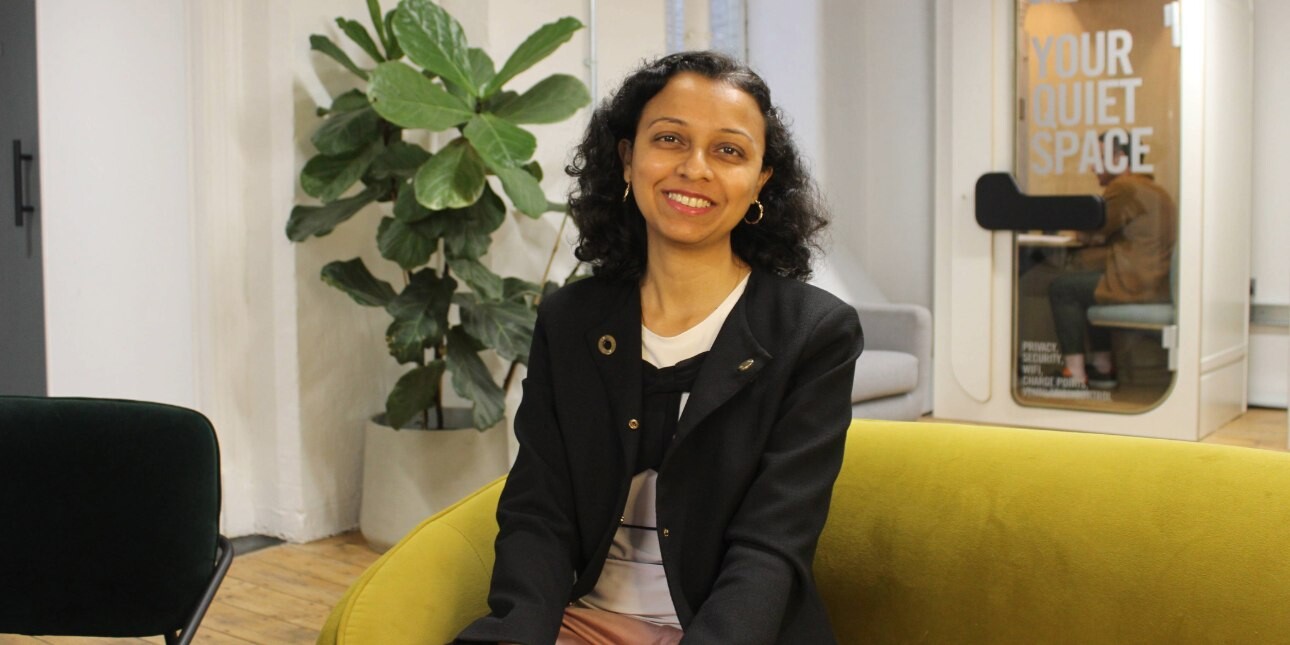 A colour portrait of Liva Emmatty sat on a mustard sofa in an office. Liva is an Asian woman with long dark hair who is wearing a black jacket over a white top and pink trousers.