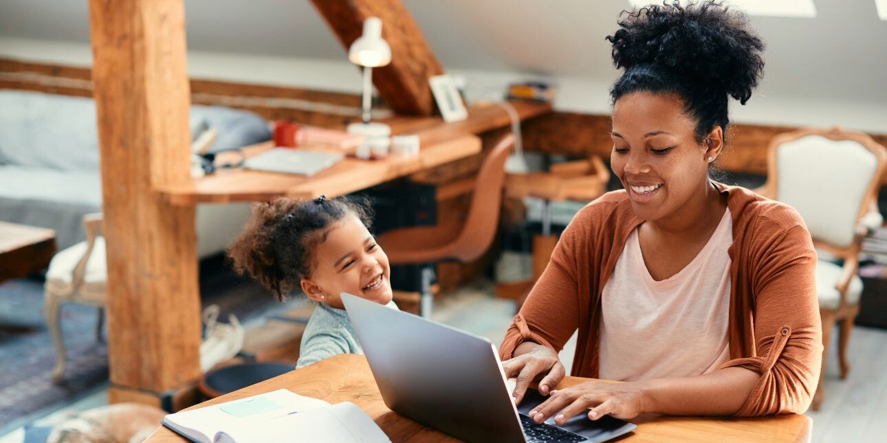 In a light filled home, a smiling Black woman wearing an orange cardigan works at her laptop as her child looks up at her smiling.
