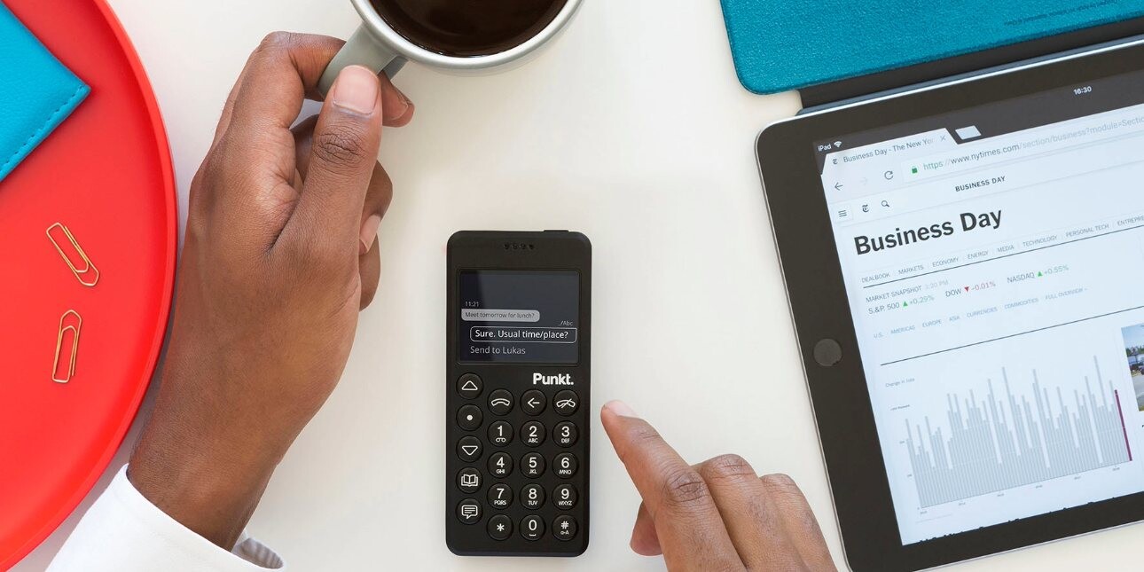 An unidentified right hand operates a Punkt device while a cup of coffee is held in the left hand. Adjacent is a tablet with a screen showing a business day calendar.
