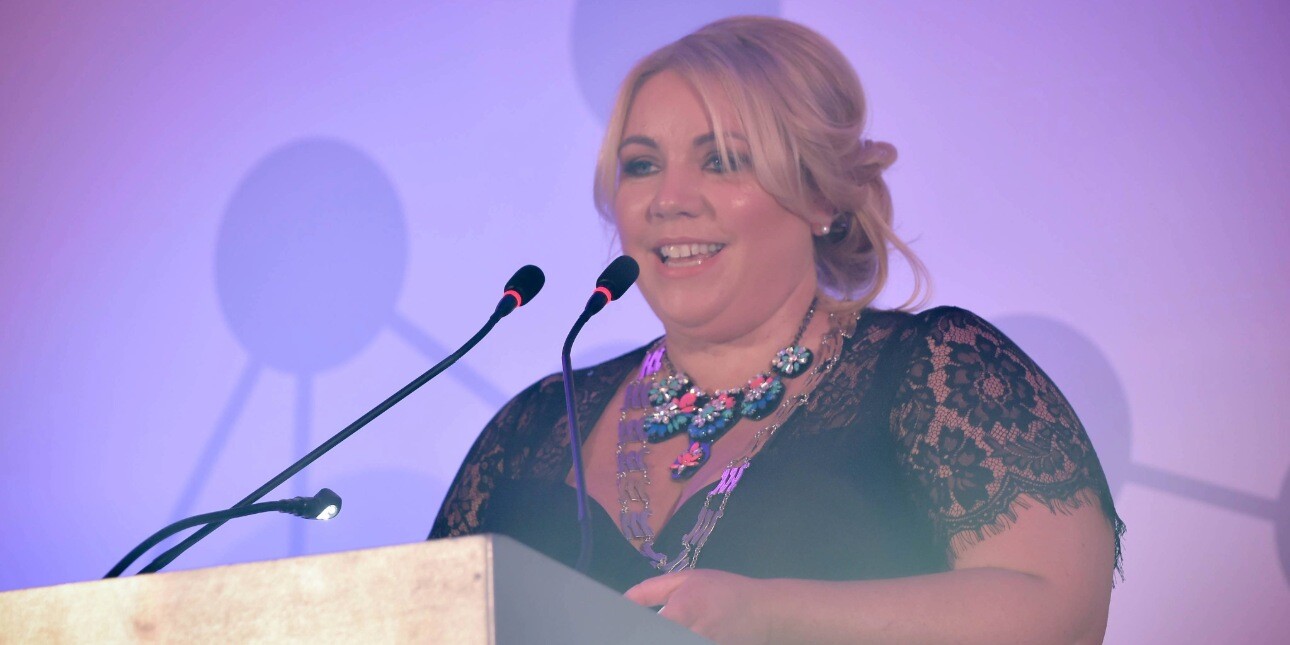 Samantha Livingstone stood at a lecturn under pink lighting. Samantha is a white woman with blond hair who is wearing a black top with laced sleeves.