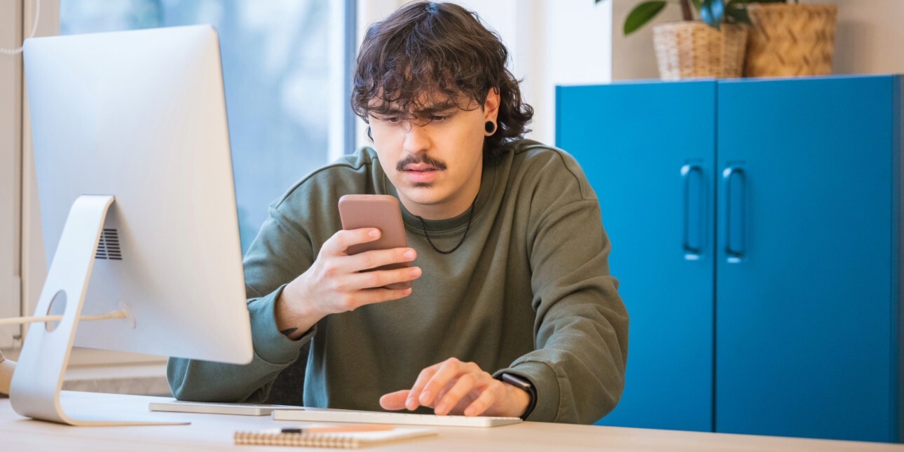 A young white man with dark hair and moustache grimaces as he stares at the mobile in his hand. He is sat in a front of a computer at a desk with a blue cabinet to the right of the image.