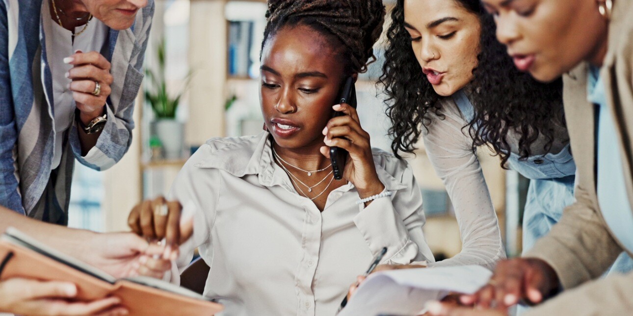 A Black business woman is sat multi-tasking as four colleagues of different genders and ethnicities try to get her attention with notebooks and documents to sign.