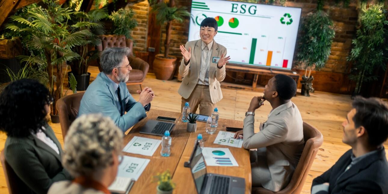 An Asian businesswoman wearing a beige suit is stood in front of a screen featuring the acronym ESG while addressessing a group of male and female colleagues sat at a board table.