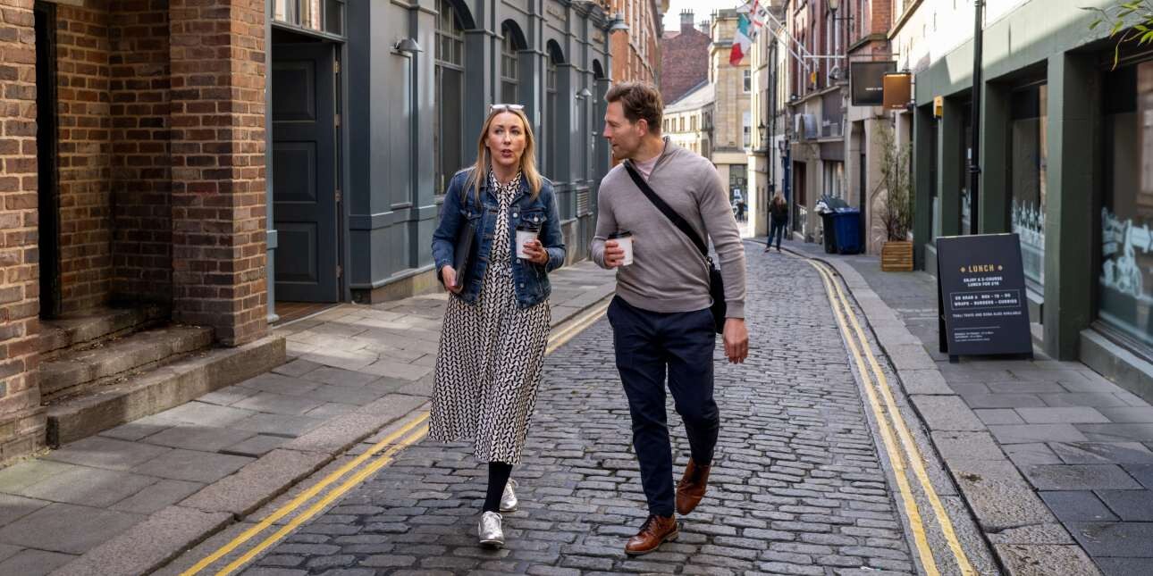 A white woman and white man in smart-casual clothing walk together along a narrow cobbled British high street while holding coffee cups.
