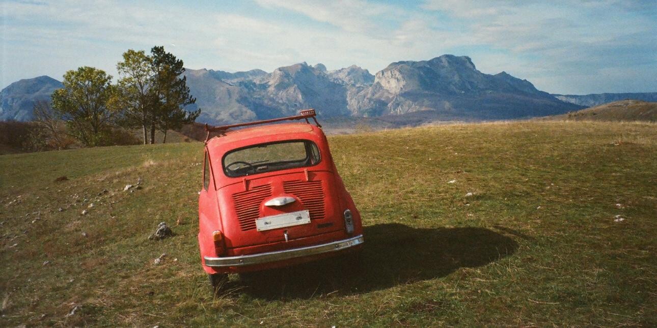 The rear of a small red vintage car parked on grass with trees and mountains in the background