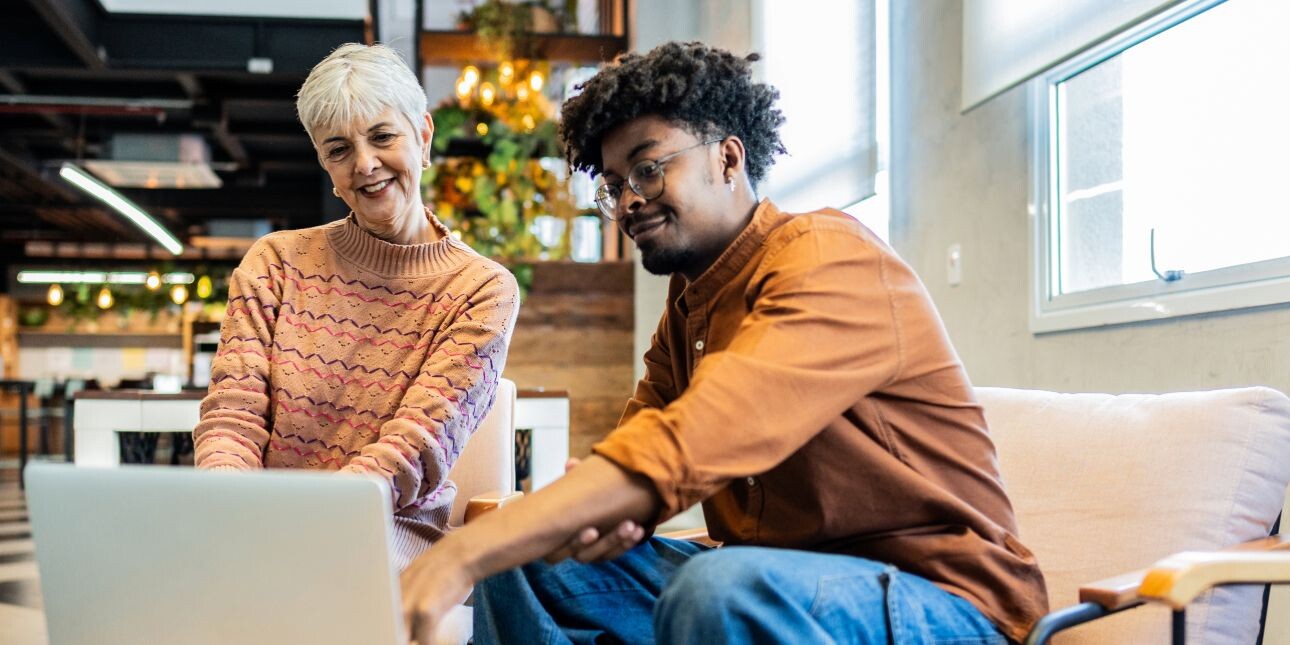 A senior white businesswoman wearing a pink patterned jumper is sat next to a Black, male middle management colleague who is wearing an orange shirt and blue jeans. They are looking at a laptop on a coffee table.