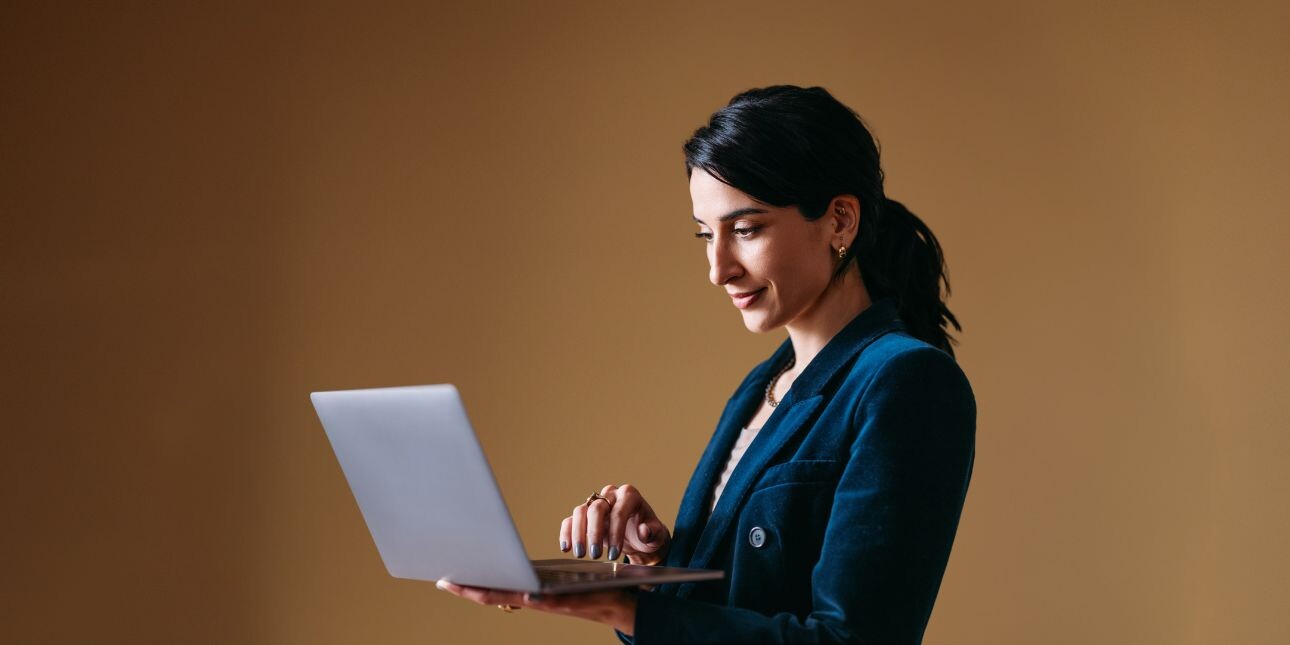 Against a brown background a business woman with dark hair and dressed in blue holds a laptop in her hand.