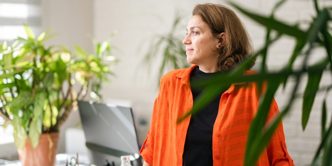 A middle-aged woman with brown hair and orange jacket over a black top looks to her right while stood in front of a desk in a light office. There are plants in the shot.