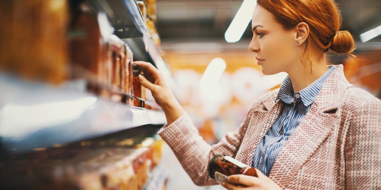 A white woman with red hair, who is wearing a plaid jacket, holds a jar of sun-dried tomatoes while browsing supermarket shelves.