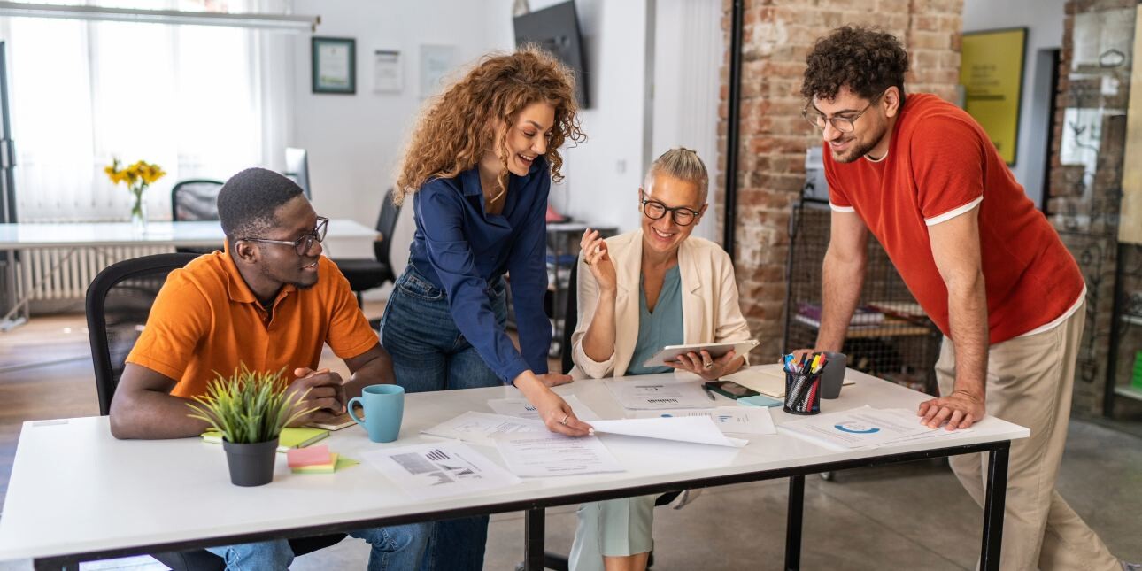 Four colleagues of different genders and ethnicities discuss paperwork on a table in a modern office.