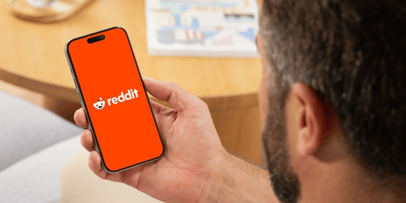 A white man holds a mobile phone featuring the reddit logo on an orange background on the screen. His head and dark hair is partially visible.
