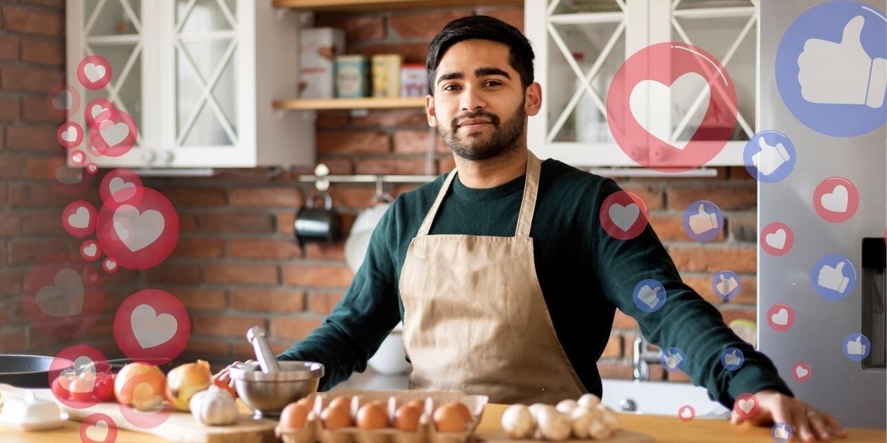 A young Asian man with short hair, who is wearing a beige apron over a green sweatshirt, stands in front of a kitchen counter preparing to cook vegetables. Semi-translucent social media hearts and thumbs up can be seen on the right hand side.