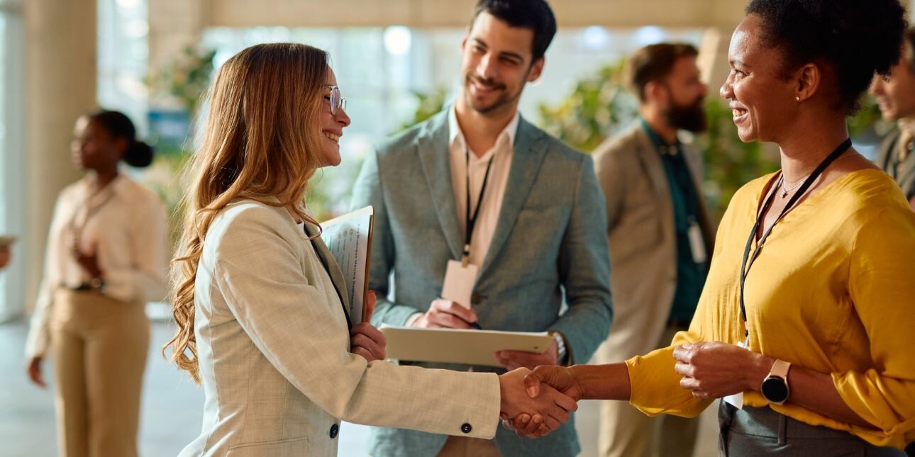 Two businesswomen shaking hands during a conference meeting, while a male colleague looks on.