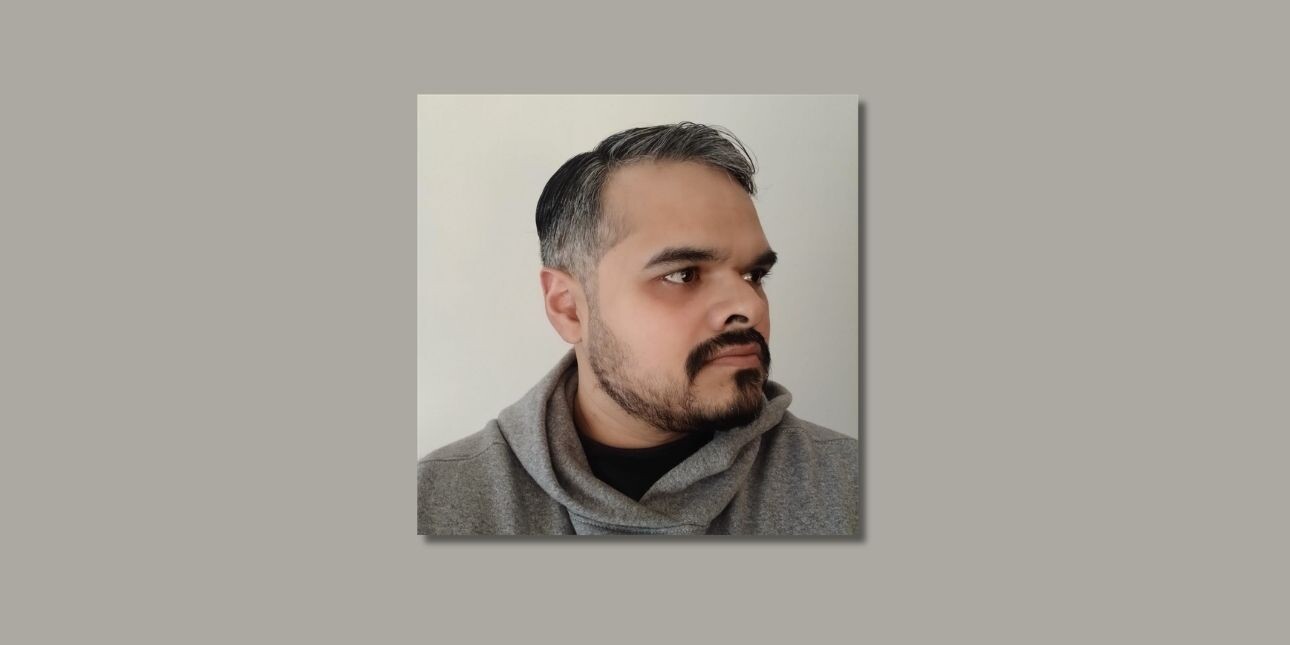 On a grey background, a colour side profile of Babar Khan Javed. Babar has short dark-grey hair and trimmed beard, and a grey hoody.